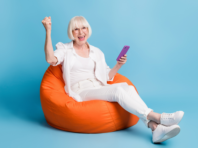 Eine fröhliche ältere Frau sitzt mit Smartphone auf einem orangefarbenen Sitzsack und jubelt begeistert.