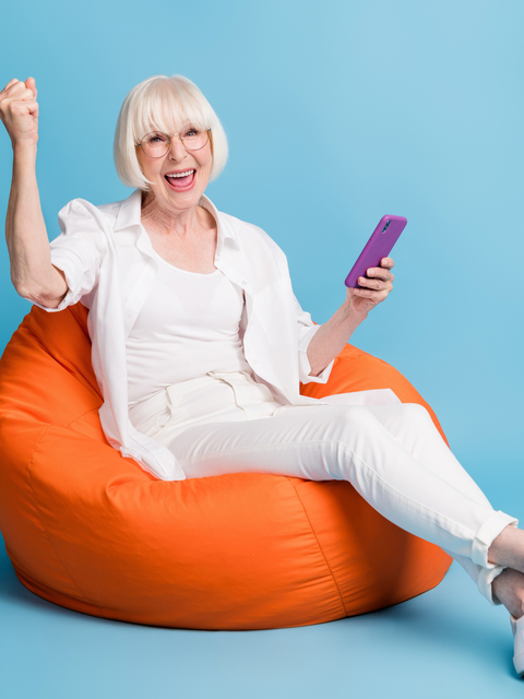 Eine fröhliche ältere Frau sitzt mit Smartphone auf einem orangefarbenen Sitzsack und jubelt begeistert.