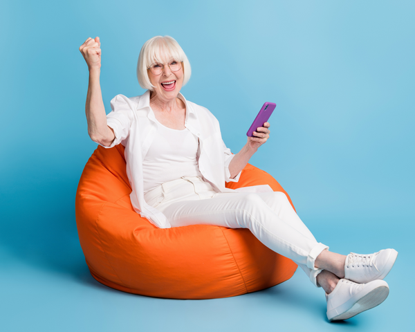 Eine fröhliche ältere Frau sitzt mit Smartphone auf einem orangefarbenen Sitzsack und jubelt begeistert.