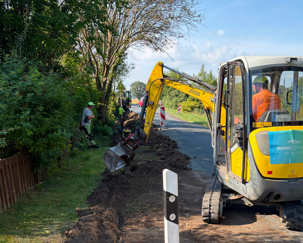 Glasfaserausbau von weserconnect: Mit einem Bagger wird entlang einer Straße ein Graben für die Verlegung der Glasfaserinfrastruktur ausgehoben. Mehrere Bauarbeiter begleiten die Arbeiten.