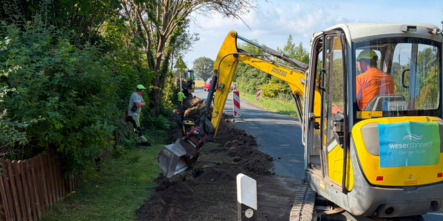 Glasfaserausbau von weserconnect: Mit einem Bagger wird entlang einer Straße ein Graben für die Verlegung der Glasfaserinfrastruktur ausgehoben. Mehrere Bauarbeiter begleiten die Arbeiten.