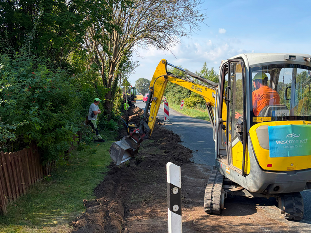 Glasfaserausbau von weserconnect: Mit einem Bagger wird entlang einer Straße ein Graben für die Verlegung der Glasfaserinfrastruktur ausgehoben. Mehrere Bauarbeiter begleiten die Arbeiten.