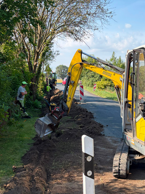 Glasfaserausbau von weserconnect: Mit einem Bagger wird entlang einer Straße ein Graben für die Verlegung der Glasfaserinfrastruktur ausgehoben. Mehrere Bauarbeiter begleiten die Arbeiten.