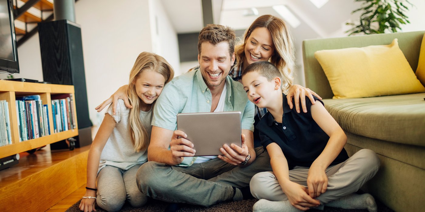 Eine Familie sitzt gemeinsam im Wohnzimmer auf dem Boden und schaut lächelnd auf ein Tablet.